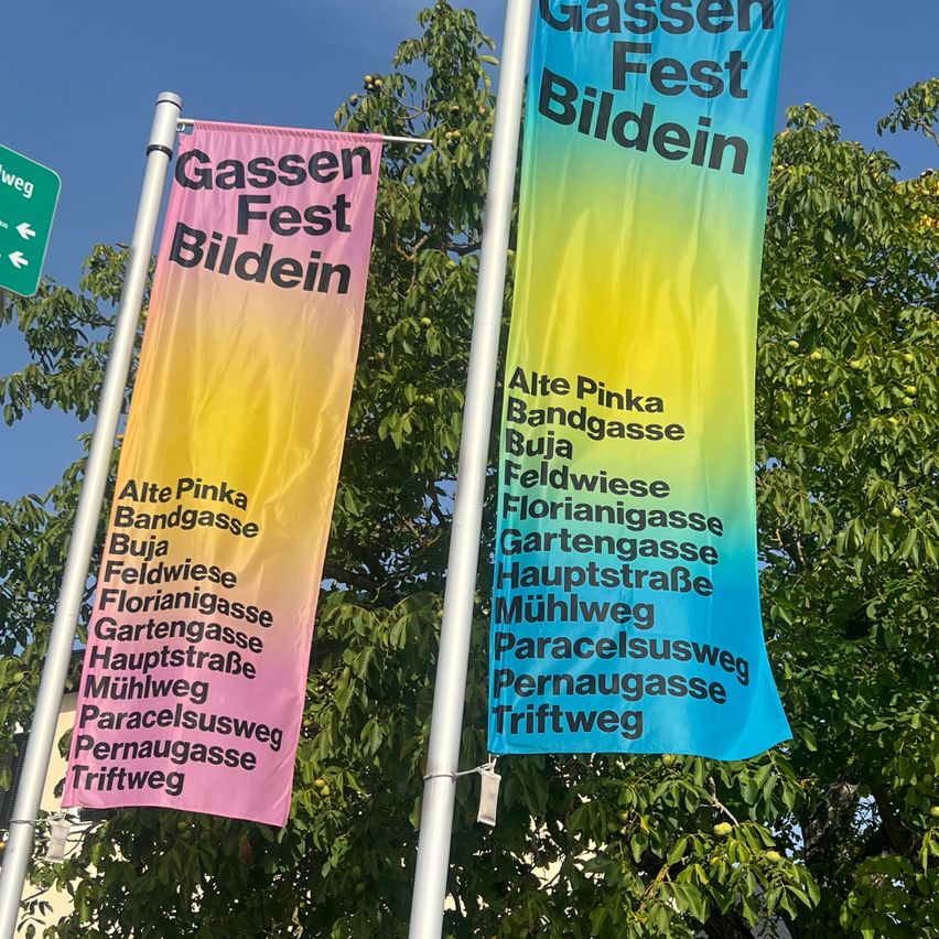 Two banners with the text Gassen Fest Bildein are displayed on poles in front of a tree. The banners are pink and blue with white text. The pink banner has the text Alte Pinka Bandgasse Buja Feldwiese Florianigasse Gartengasse Hauptstraße Mühlweg Paracelsusweg Pernaugasse Triftweg. The blue banner has the text Alte Pinka Bandgasse Buja Feldwiese Florianigasse Gartengasse Hauptstraße Mühlweg Paracelsusweg Pernaugasse Triftweg.