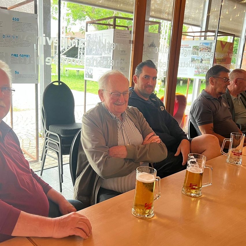 Fünf Männer sitzen mit Bierkrügen um einen Tisch in einem Raum mit Glaswänden und Plakaten.