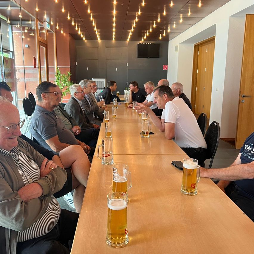 Eine Gruppe älterer Männer sitzt an einem langen Tisch in einer Bar, mit Bierkrügen. Der Raum hat eine warme Atmosphäre mit gedämpfter Beleuchtung und Holztüren.