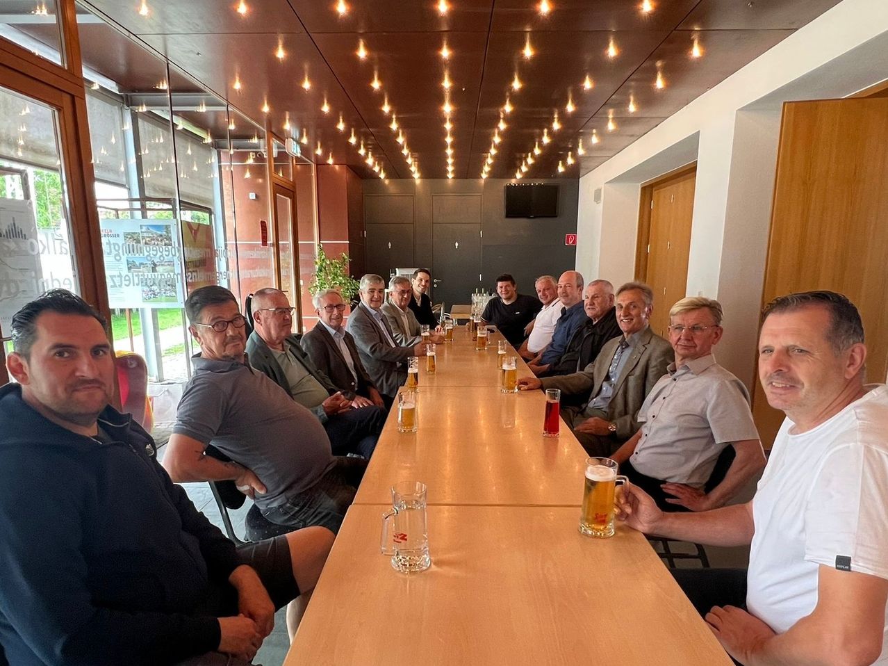 Eine Gruppe älterer Männer sitzt um einen langen Tisch in einem Restaurant, jeder mit einem Bier. Sie lächeln, manche halten ihre Hände hinter dem Rücken.