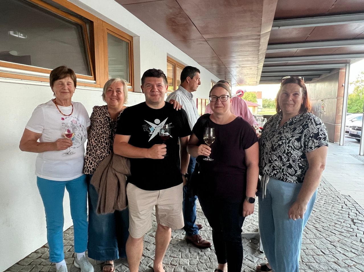 A group of five adults, two women and three men, are standing together in a hallway, smiling and holding wine glasses. The man in the center is wearing a black t-shirt with a white design.
