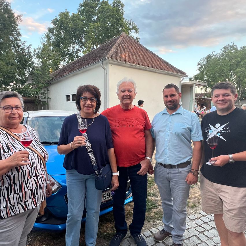 Five people are standing in front of a blue car and a white house. They are all holding wine glasses and smiling. The man in the middle is wearing a red shirt. The woman on the left is wearing glasses and a necklace. The man on the right is wearing a watch.