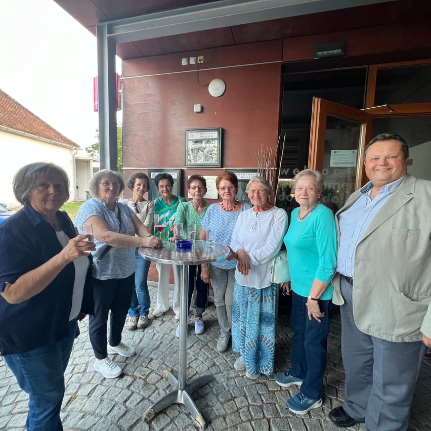 A group of elderly women and men stand around a table with glasses and a bowl, smiling for a photo in front of a building.
