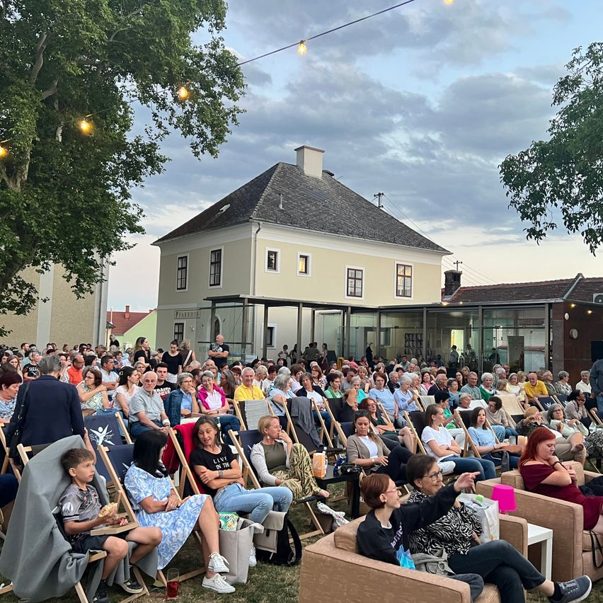 A large crowd of people are seated on chairs in an outdoor area, possibly for an event. The chairs are arranged in rows, and some people are standing. The area is surrounded by trees, and a building with glass windows is in the background. The sky is cloudy.