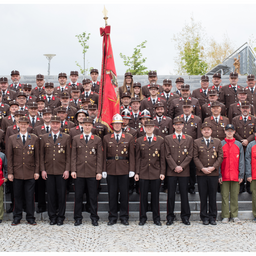 Eine Gruppe uniformierter Personen, einige mit Mützen, steht in Reihen auf Treppen. Ein Mann in der Mitte hält eine Flagge. Hinter ihnen stehen einige Bäume und ein Gebäude.