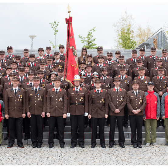 Eine Gruppe uniformierter Personen, einige mit Mützen, steht in Reihen auf Treppen. Ein Mann in der Mitte hält eine Flagge. Hinter ihnen stehen einige Bäume und ein Gebäude.
