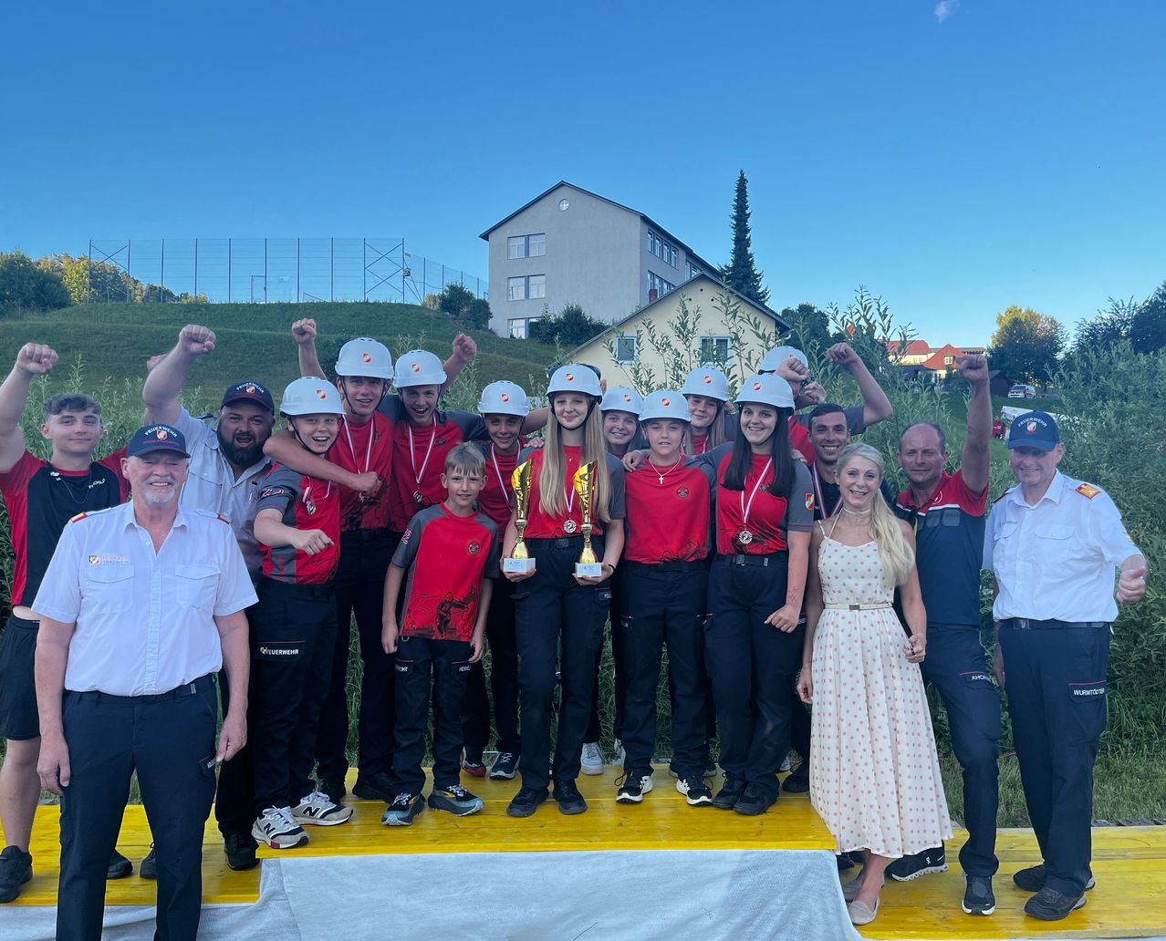 Eine Gruppe junger Menschen in roten und weißen Helmen feiert mit Trophäen auf einer gelben Plattform. Sie stehen neben einigen Erwachsenen im Hintergrund.