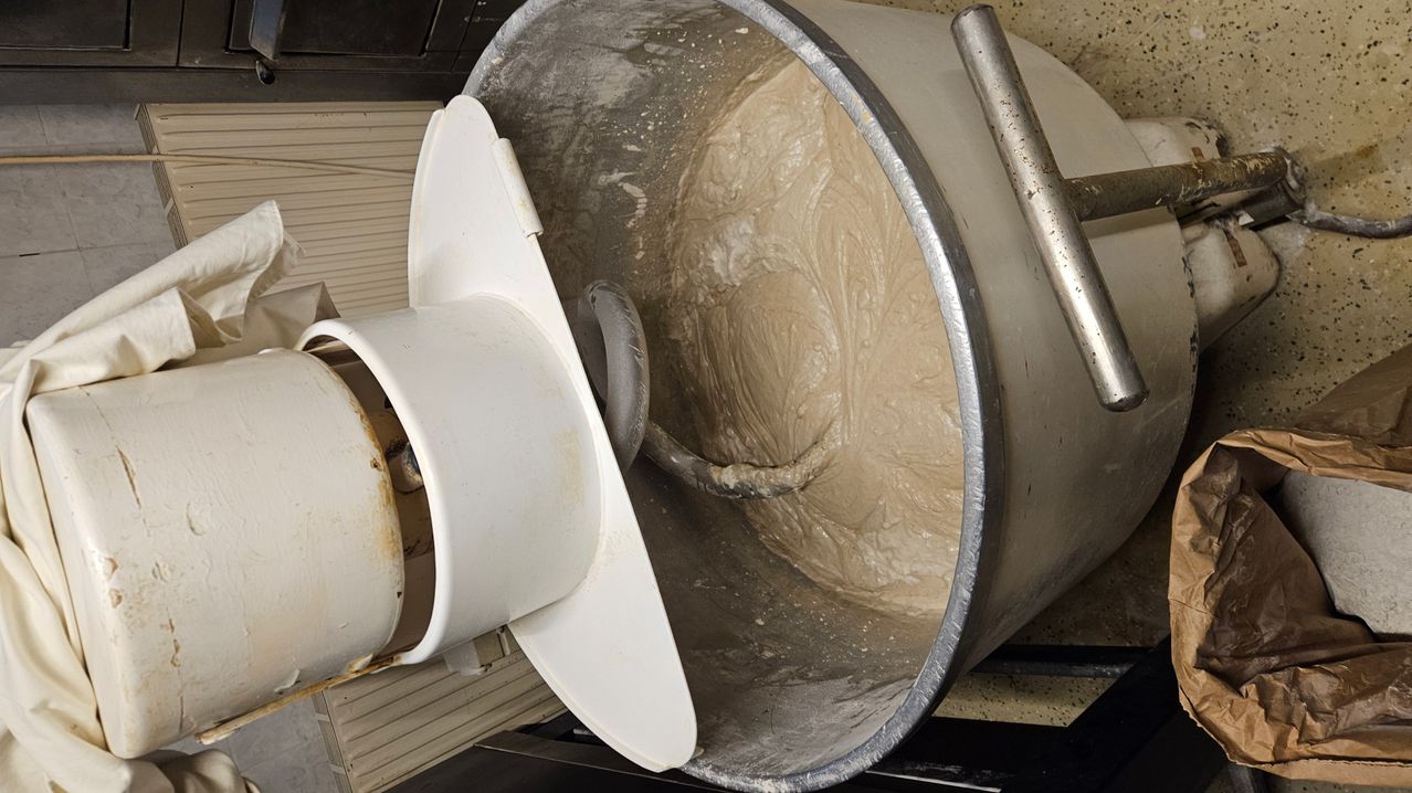 Ein großer Edelstahlmixer mit weißem Deckel mischt einen hellbraunen Teig. Der Mixer steht auf einem gefliesten Boden.