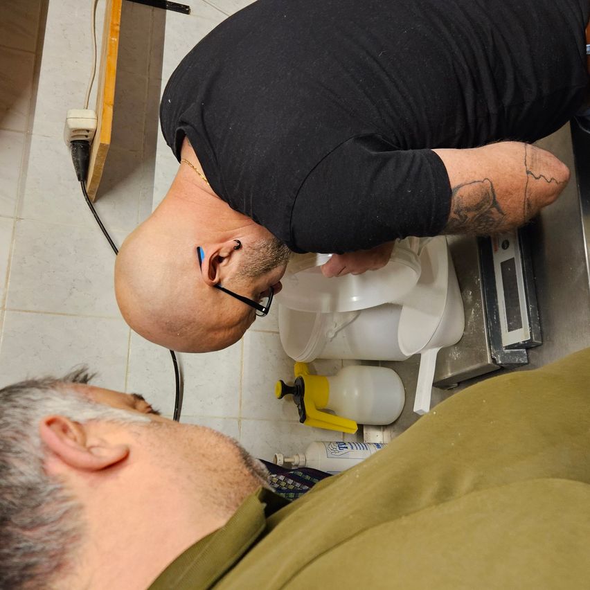 Ein Mann mit einer Tätowierung am Arm beugt sich über einen Behälter, während ein anderer Mann mit einer Maske im Gesicht auf dem Boden liegt.