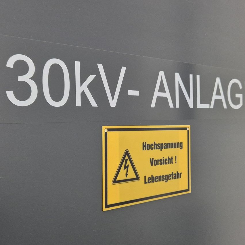 Ein graues Schild mit weißem Text steht '30kV-ANLAGE'. Darunter befindet sich ein gelbes Schild mit Blitzsymbol, das vor 'Hochspannung' warnt. Es steht 'Vorsicht! Lebensgefahr'.