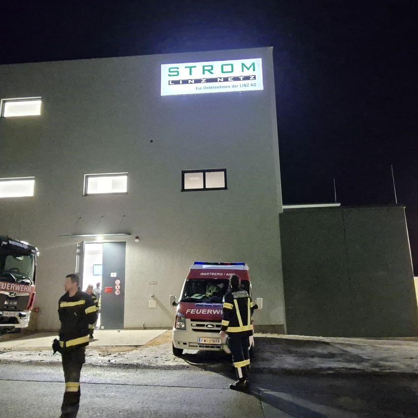 Zwei Feuerwehrleute stehen vor einem Krankenwagen vor einem Gebäude mit einem Strom Linz Netz-Schild bei Nacht.