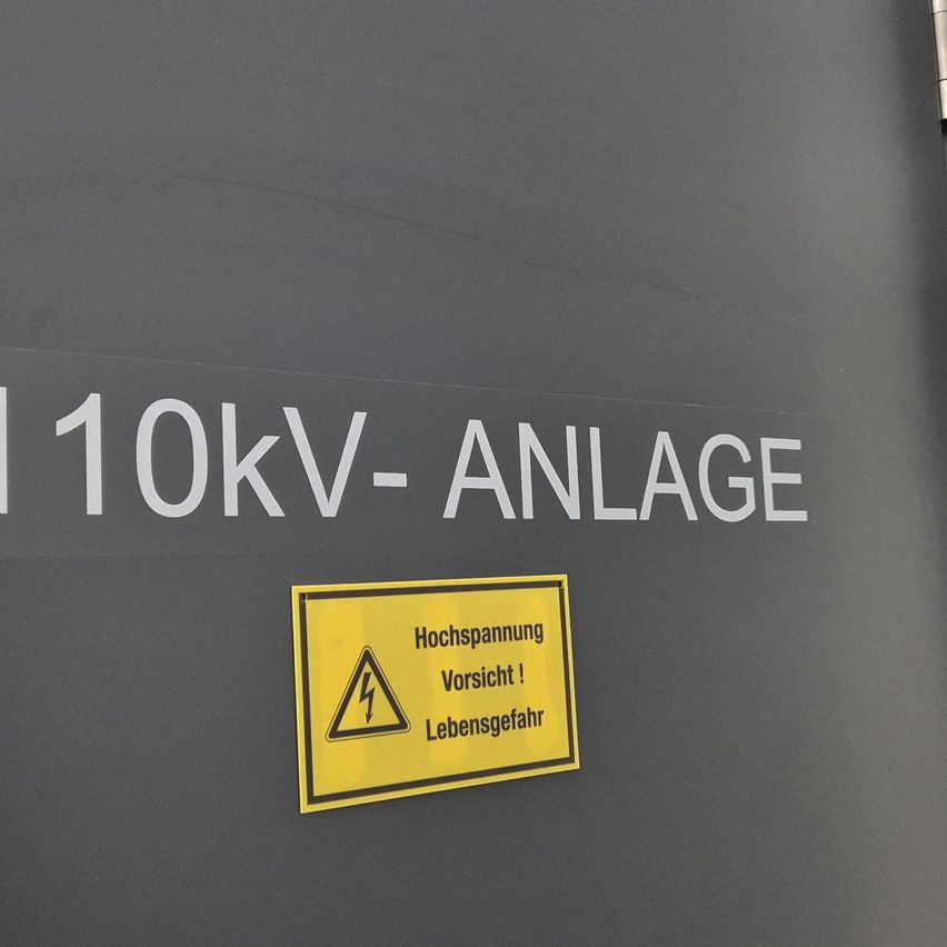 Eine graue Wand mit weißem Text, der '110kV-ANLAGE' lautet. Darunter befindet sich ein gelbes Schild mit einem Blitzsymbol, das vor hoher Spannung und Lebensgefahr warnt.