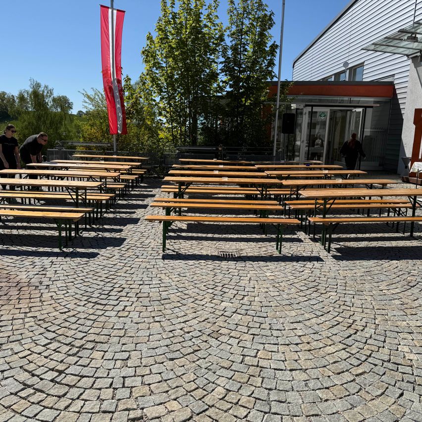 Eine Flagge steht vor einem Gebäude mit hölzernen Bänken und Tischen, die in einem Kreis angeordnet sind. Einige Leute richten das Setup ein. Der Himmel ist klar und blau.