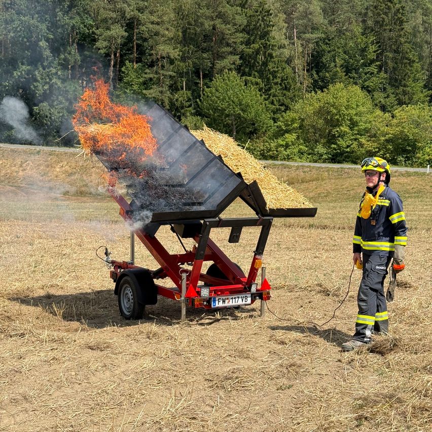 A firefighter in full gear stands next to a red trailer with a fire-extinguishing device burning hay on top, set against a backdrop of a field and trees.