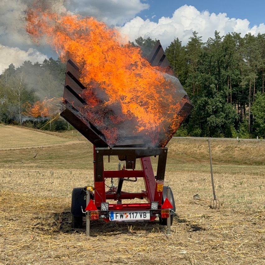 A red trailer with a license plate FW 117 VB is on fire in a field with dry grass.