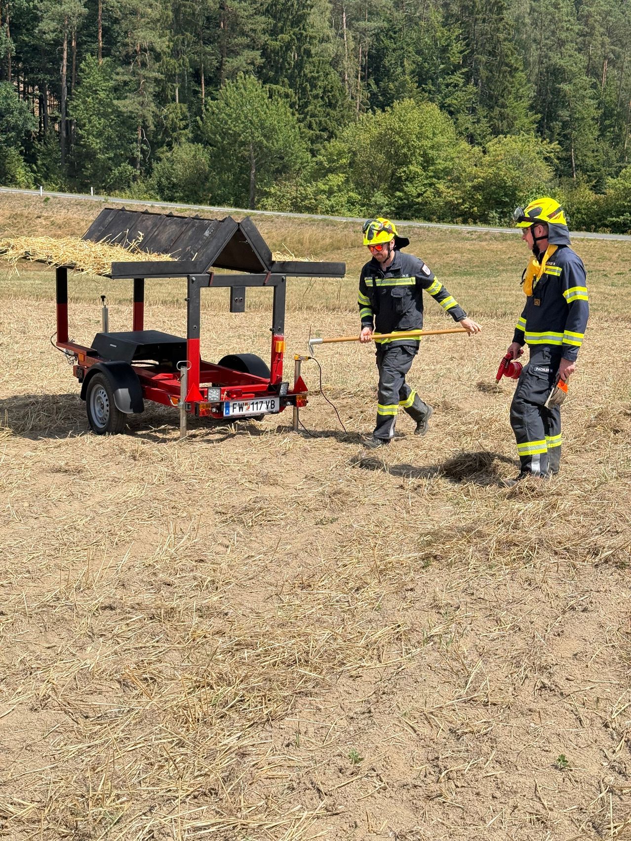 Two firefighters in full gear are walking towards a trailer with a hay roof in a dry field.