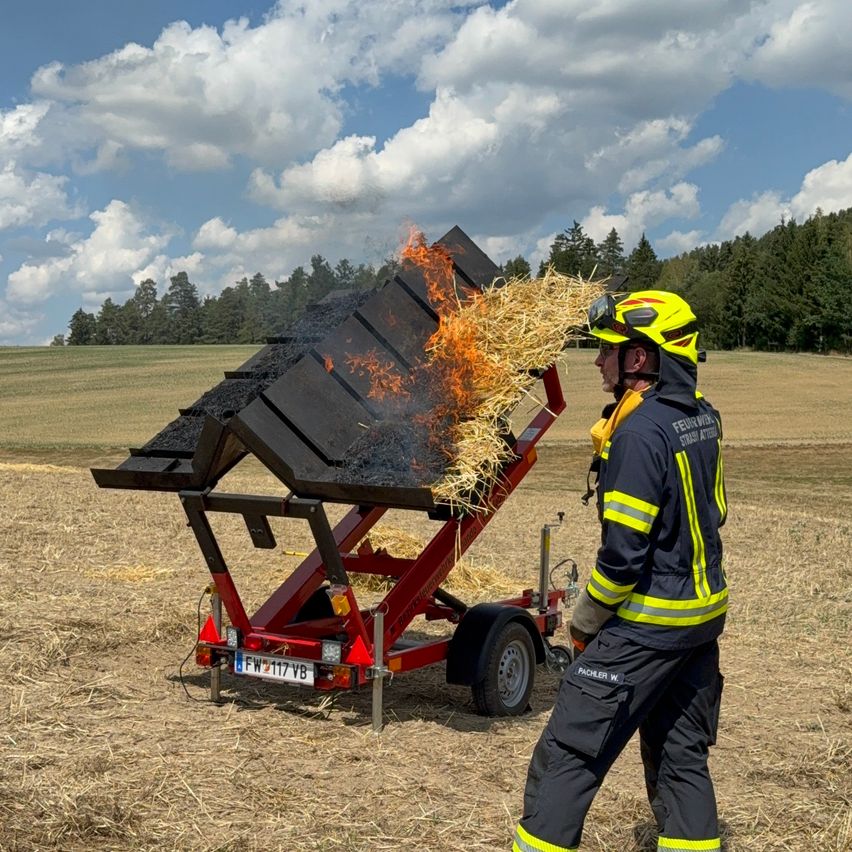 A firefighter in full gear stands next to a fire-fighting trailer with hay on fire in a field.