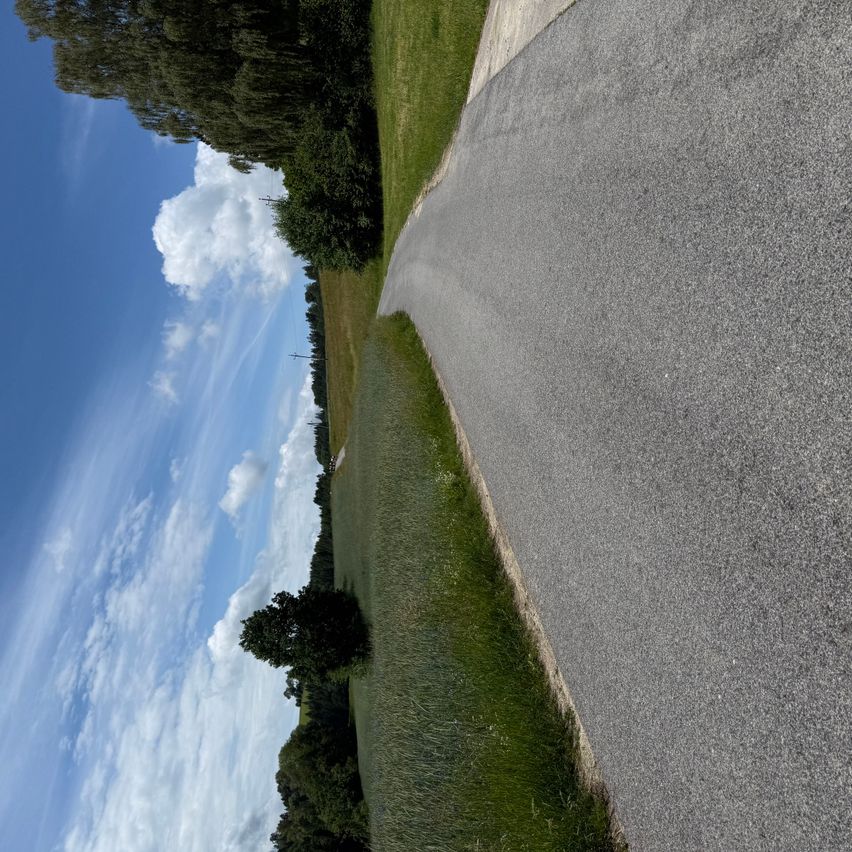 An aerial view of a road with gravel, surrounded by green grass and trees under a blue sky with clouds.