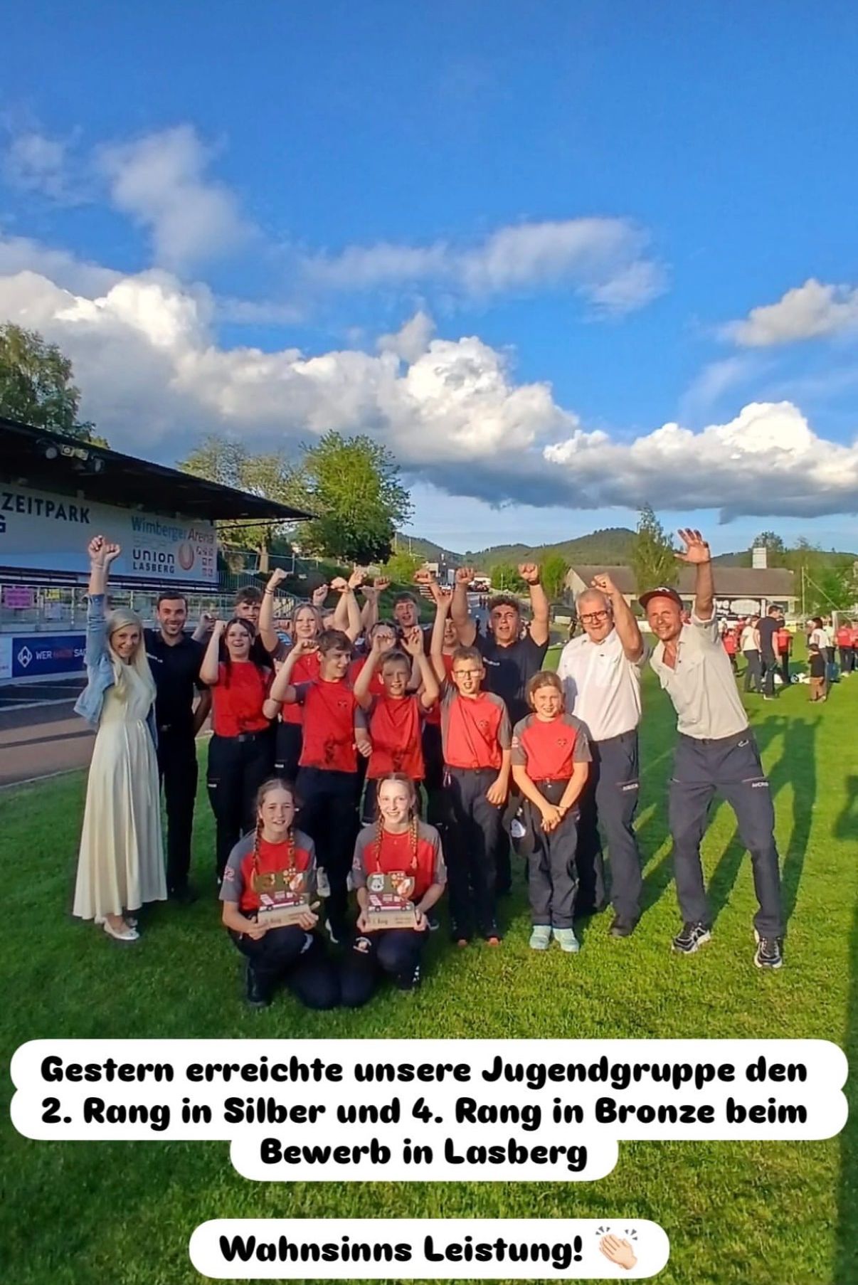 A group of people stands on a grassy field, smiling, holding trophies. Some wear red shirts, while others wear different clothes. A woman in a white dress stands on the left. A banner reads 'Wimberger Arena'.