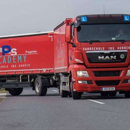 Ein roter MAN-Lkw mit der Aufschrift 'Fahrschule Ing. Aubock' fährt auf einer Straße mit einem Grasstreifen am Straßenrand.