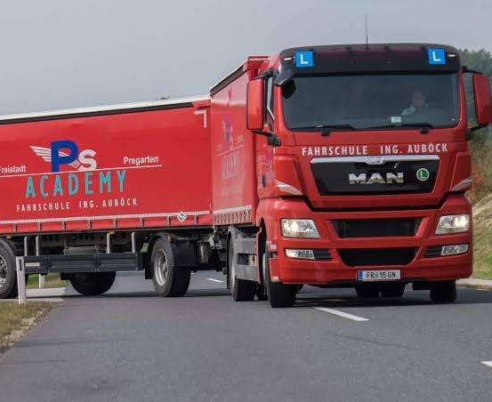 Ein roter MAN-Lkw mit der Aufschrift 'Fahrschule Ing. Aubock' fährt auf einer Straße mit einem Grasstreifen am Straßenrand.