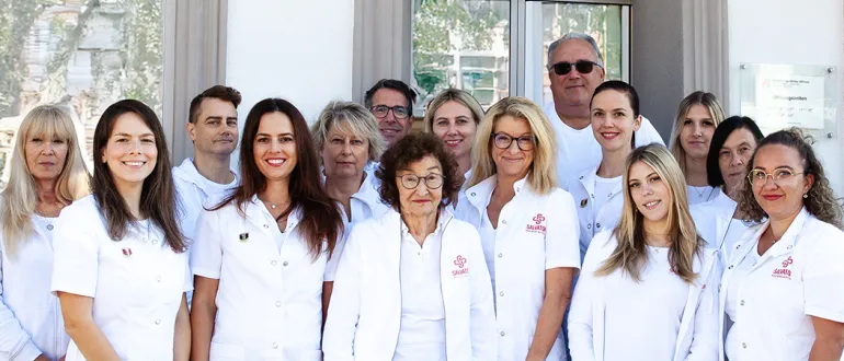 Eine Gruppe medizinischer Fachkräfte, darunter Frauen in weißen Kitteln und Brillen, steht in einer Reihe vor einem Gebäude mit Glasfenstern und einem reflektierenden Schild.