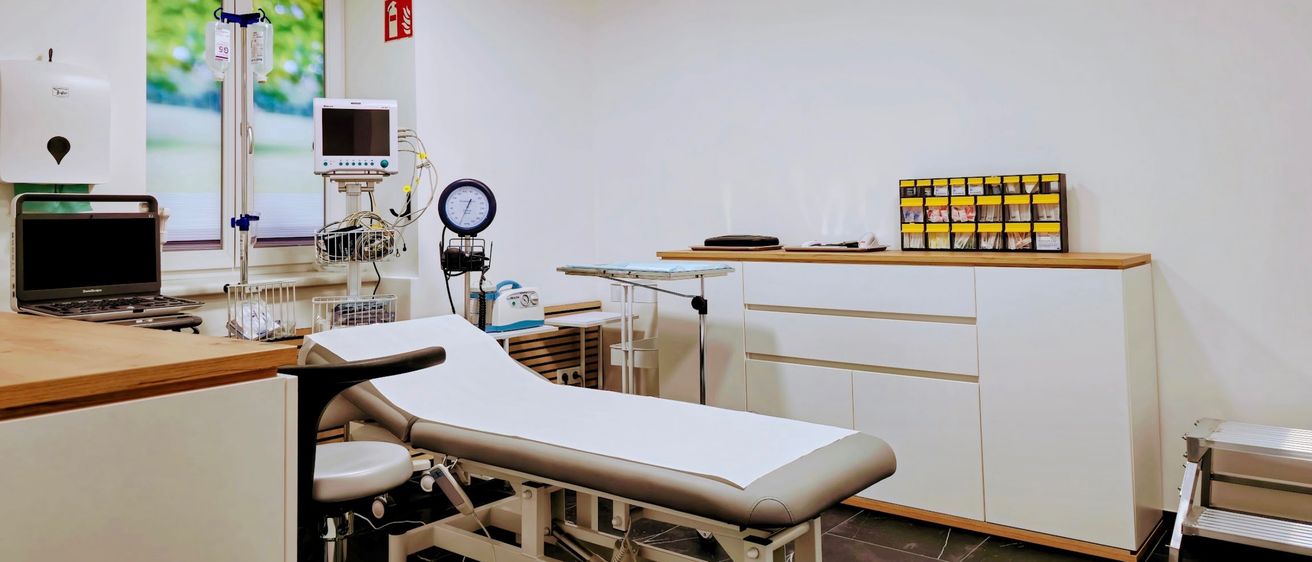 Ein leerer medizinischer Untersuchungsraum mit einem Monitor, einem Sphygmomanometer, einem Stuhl und einem Tisch. An der Wand ist ein Feuerlöscher angebracht.