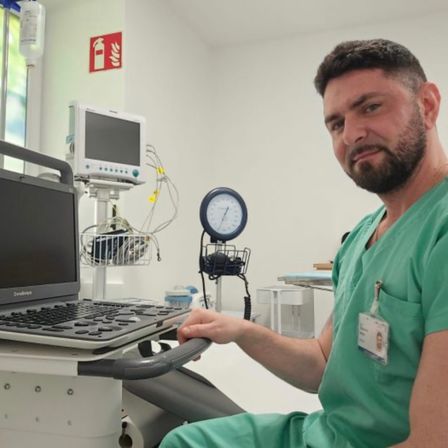 Ein Mann in einem grünen Krankenhausuniform sitzt vor einem medizinischen Monitor und einer Tastatur. Hinter ihm ist ein Feuerlöscher an der Wand montiert.