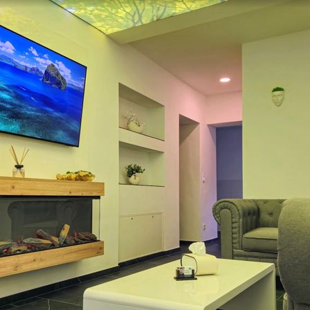 Ein elegantes Wohnzimmer mit einem großen Flachbildfernseher, einem Kamin und einem weißen Sofa. Auf dem Tisch befinden sich ein Taschentuchspender und eine Karte.