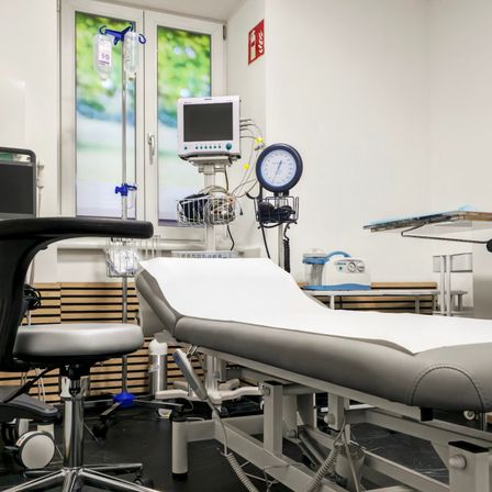 Ein medizinischer Raum mit einem Untersuchungstisch, einem Monitor und verschiedenen medizinischen Geräten. Der Raum hat weiße Wände, Glasfenster und ein Feuerlöscher-Schild.