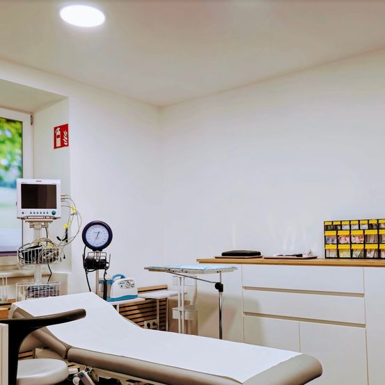 Ein Untersuchungsraum in einer Klinik mit weißen Wänden, Decke und Boden. Er enthält ein Krankenhausbett, einen Monitor, ein Feuerlöscher-Schild und einen Schrank mit verschiedenen medizinischen Utensilien.
