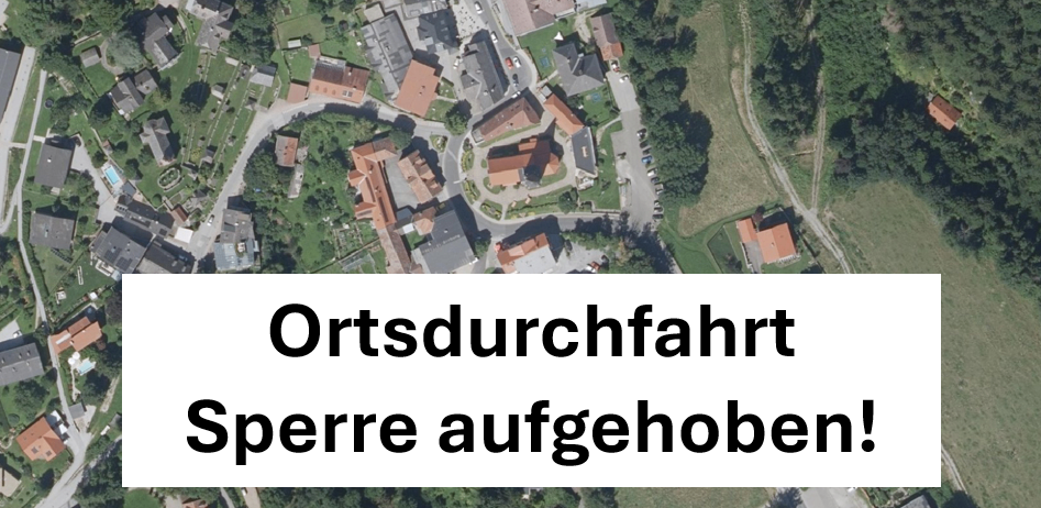 Luftaufnahme eines kleinen Ortes mit Wohngebäuden, Bäumen und einem Weg. Der Ort ist von Grün umgeben. Der Text 'Ortsdurchfahrt Sperre aufgehoben!' wird auf einem weißen Hintergrund angezeigt.