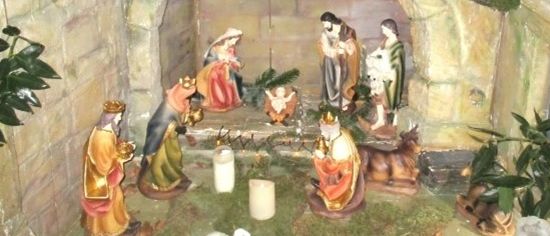Eine Krippe mit Statuen von Jesus, Maria, Josef und den drei Weisen aus dem Morgenland. Ein Baby Jesus liegt in einer Krippe, neben ihm ein kleines Lamm. Kerzen umgeben die Szene.