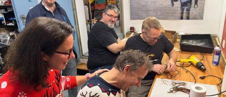 Mehrere Männer arbeiten in einem Raum an einem Projekt. Ein Mann im Rentierpullover lächelt in die Kamera. Ein anderer Mann benutzt einen Lötkolben. An der Wand steht ein großer Roboter.