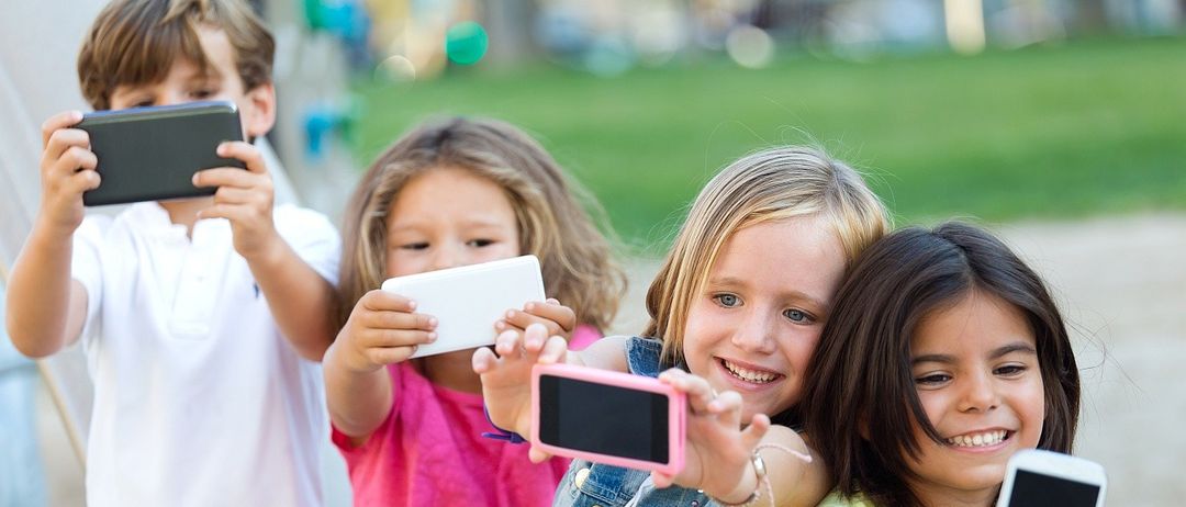 Vier Kinder, die lächeln und Handys halten, stehen in einem Außenbereich, vermutlich einem Park. Das Mädchen in der Mitte hält ein rosa Handy.