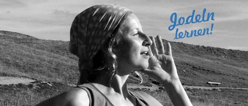 A woman in a sleeveless shirt and bandana stands in a grassy field with mountains in the background. She looks up with her hand near her face. The text 'Jodeln lernen!' is written in blue.
