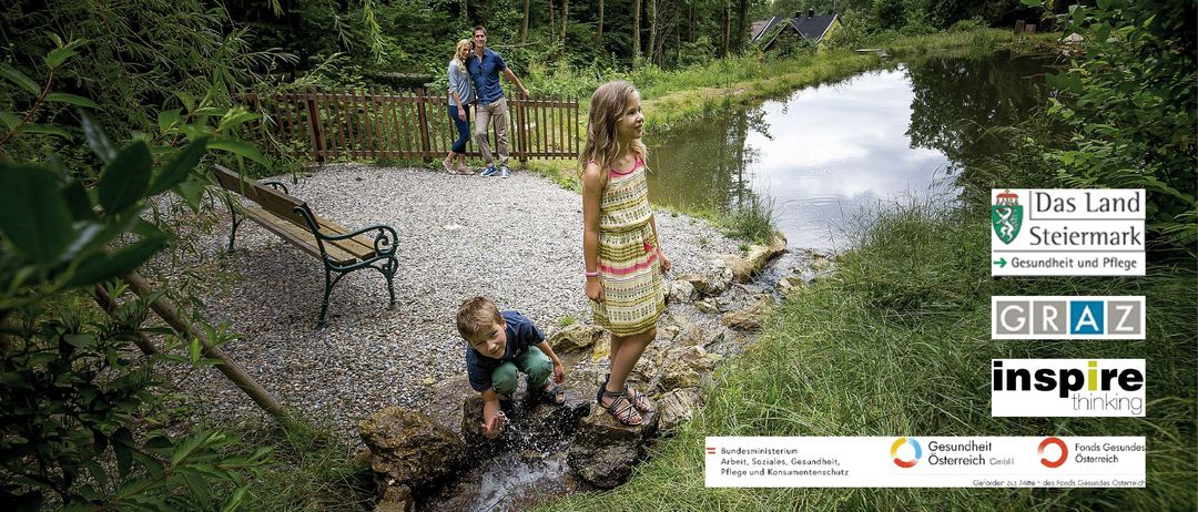 Eine Familie am Teich, ein Junge spielt mit Wasser, ein Mädchen steht daneben, ein Paar steht auf Kies, Bäume und Häuser im Hintergrund.