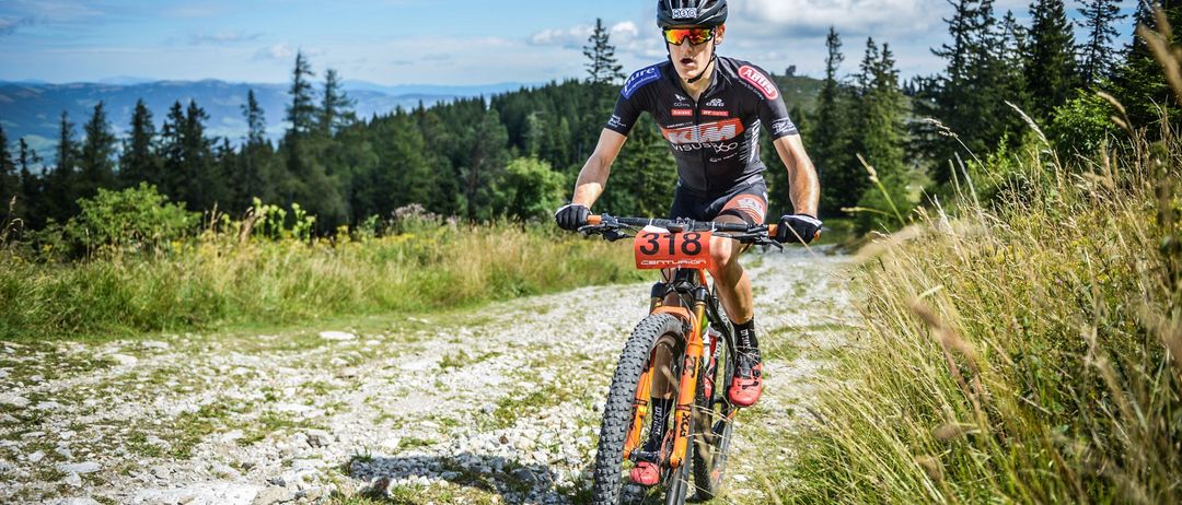Ein Radfahrer bei einem Mountainbike-Rennen, mit Helm und Radbekleidung, fährt auf einem felsigen Pfad durch einen Wald. Die Nummer 318 ist auf der Vorderseite des Fahrrads angebracht.