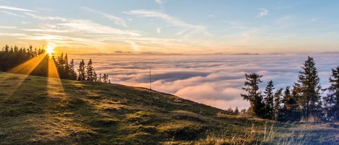 Sonnenuntergang über einer bergigen Landschaft mit Wolken darunter und Bäumen auf dem Hügel, die Sonnenstrahlen beleuchten die Szene.