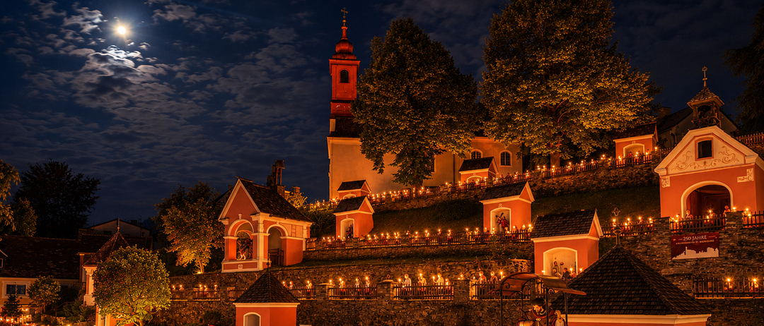 Eine Kirche, die mit Lichtern geschmückt und eine Statue im Vordergrund ist, umgeben von Menschen, die bei Nacht Kerzen halten.