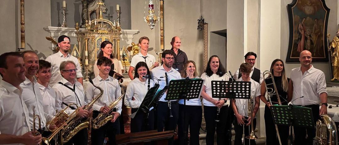 Eine Band posiert für ein Foto in einer Kirche, mit Musikern, die Instrumente wie Saxophone und Trompeten halten, und anderen, die mit Posaunen stehen. Hinter ihnen sind eine religiöse Statue und Kerzen ausgestellt.