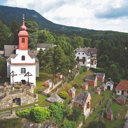 Ein Dorf mit einer Kirche, umgeben von Bäumen und Häusern, hat einen Steinzaun und kleine Kapellen. Berge sind in der Ferne unter einem bewölkten Himmel.