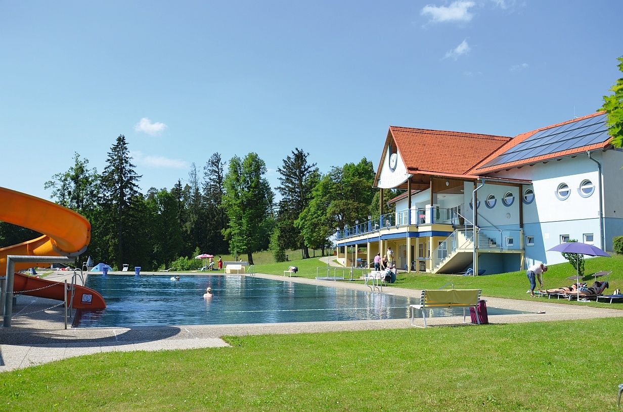Ein Freibadbereich mit einem großen Pool und einem Gebäude mit rotem Dach. Mehrere Menschen schwimmen und entspannen sich am Pool.