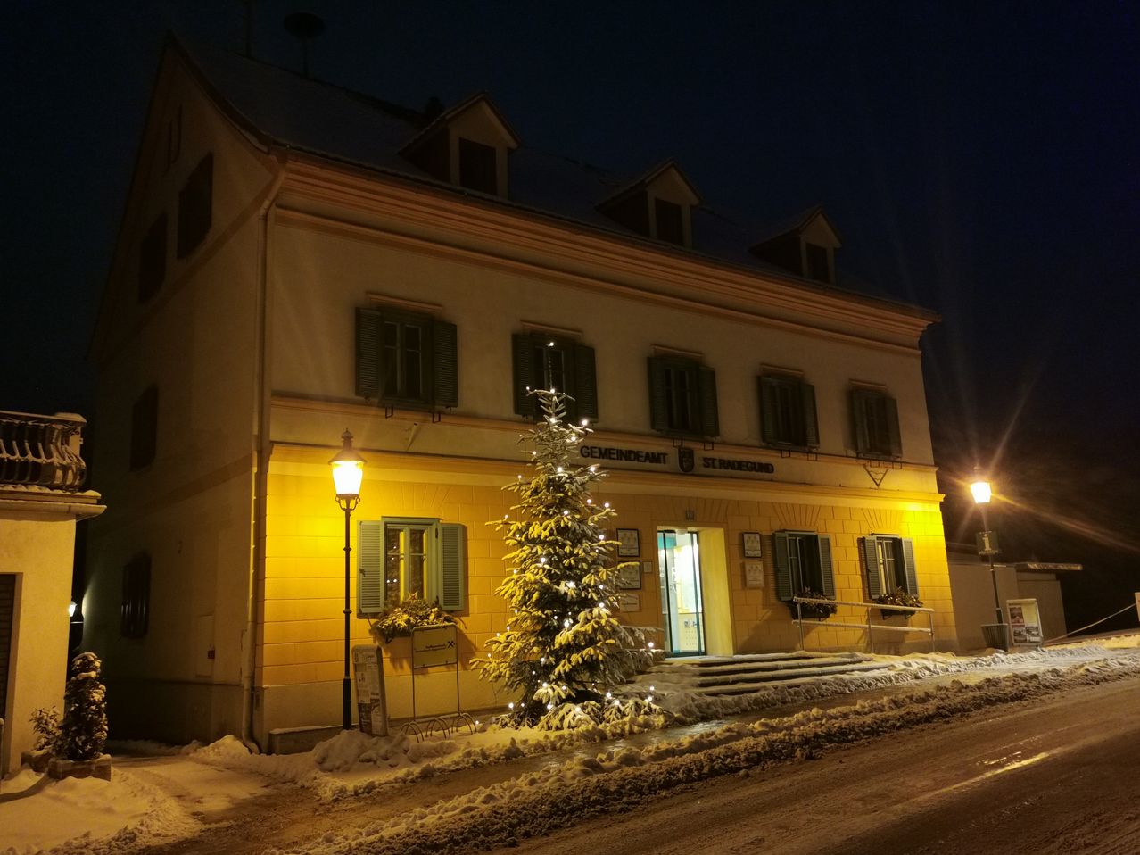 Ein gelbes Gebäude mit mehreren Fenstern, einem beleuchteten Weihnachtsbaum und Straßenlaternen davor in der Nacht.
