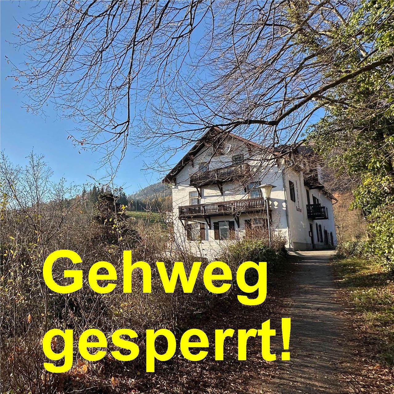 Ein weißes Haus mit Balkonen und einem Laternenpfahl steht auf einem von Bäumen und Büschen umgebenen Pfad. Das Bild zeigt den Text 'Gehweg gesperrt!' in Gelb. Im Hintergrund sind Berge und ein klarer blauer Himmel zu sehen.