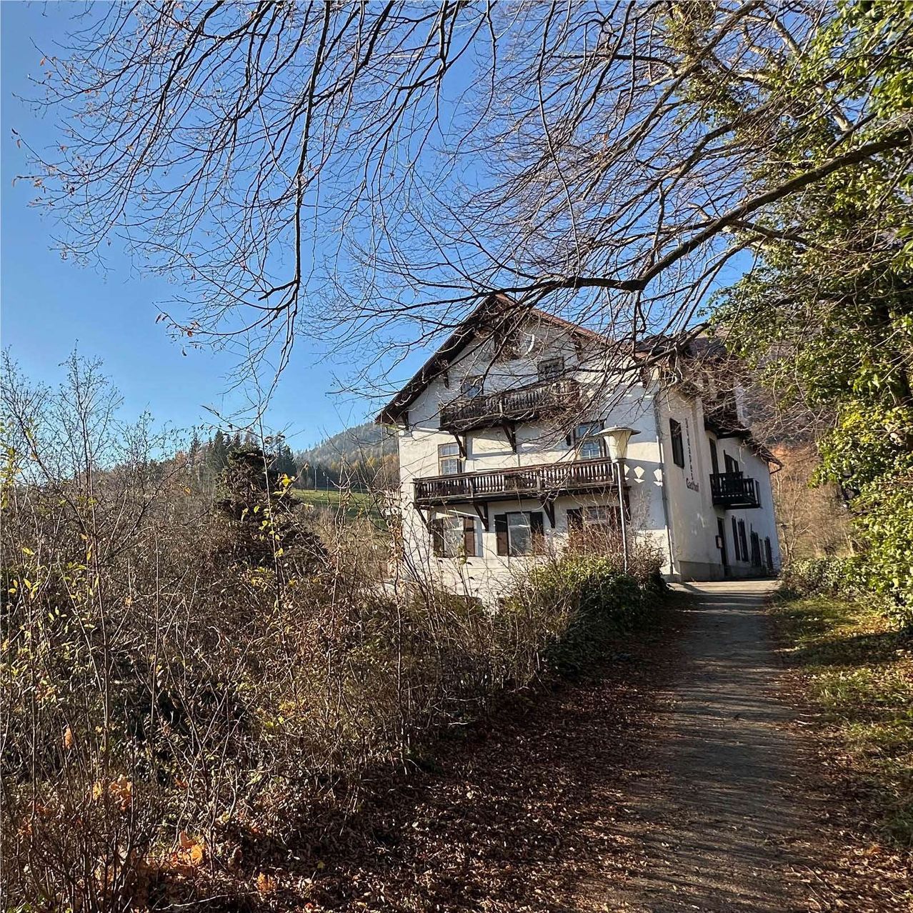 Ein weißes Haus mit Holzbalkonen ist von Grün umgeben. Ein Weg führt unter einem blauen Himmel zum Haus.