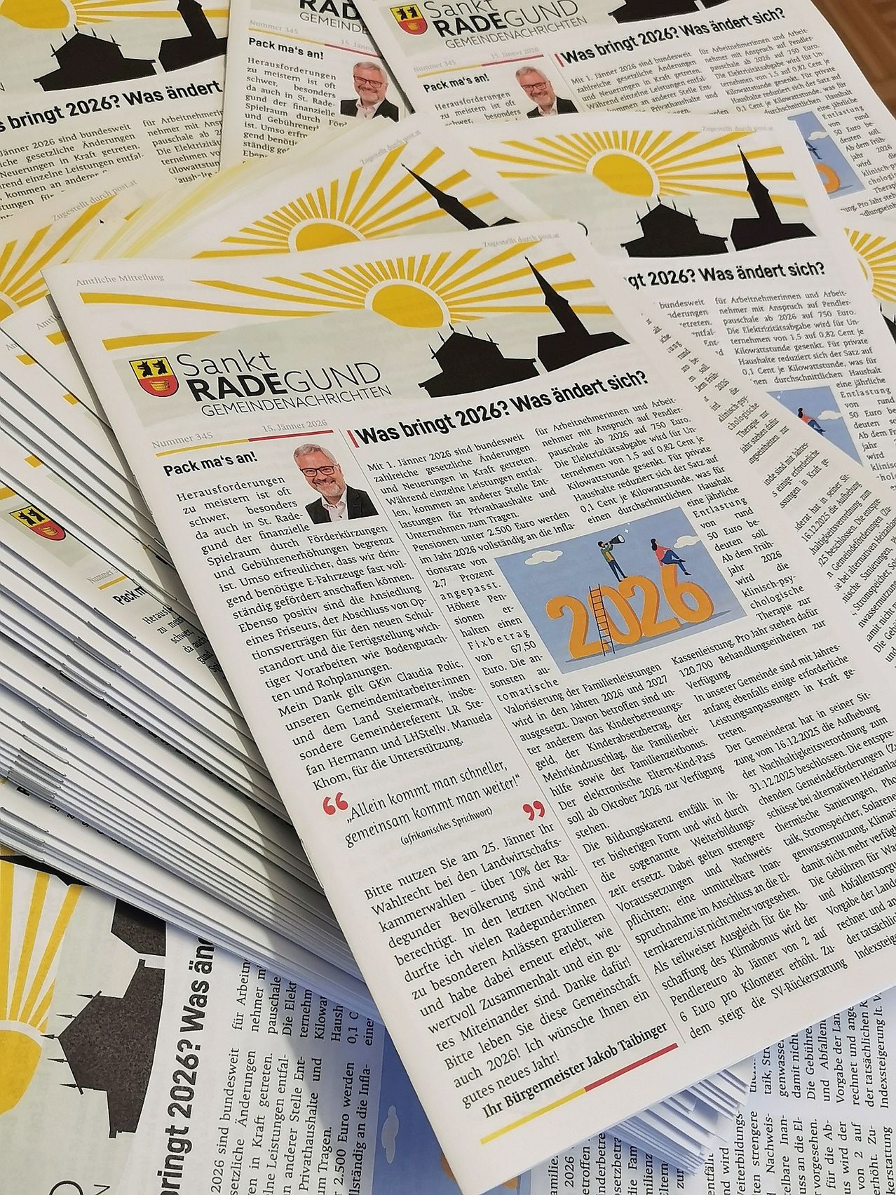 Ein Stapel gedruckter Papiere mit dem Titel 'Sankt Radegund Gemeindenachrichten' enthält einen Artikel mit der Überschrift 'Was bringt 2026?' Ein Cartoonbild einer Leiter und des Jahres 2026 ist ebenfalls vorhanden.