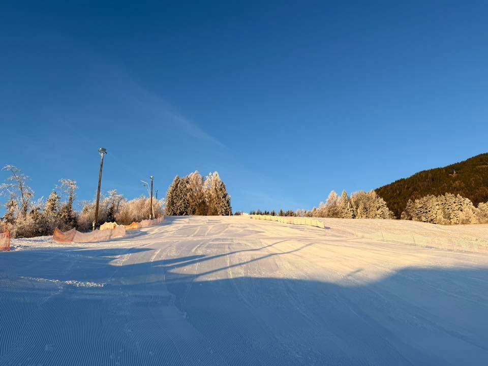 Ein Skihang unter einem klaren blauen Himmel, mit schneebedecktem Boden und Bäumen, die mit Frost bedeckt sind. Straßenlaternen stehen am Rande.