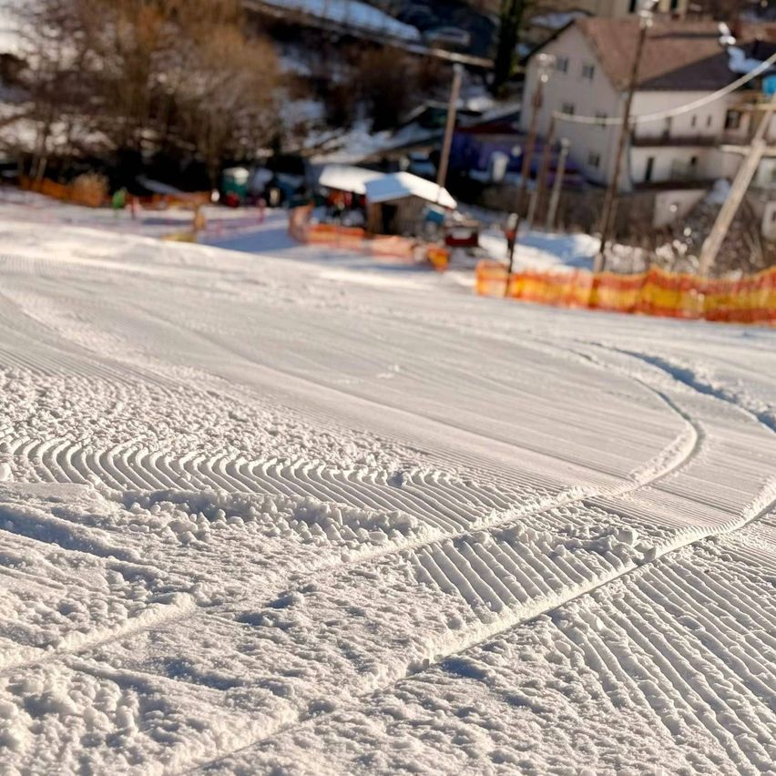 Eine verschneite Landschaft mit Reifenspuren im Schnee. Im Hintergrund sind Häuser und Bäume, die ebenfalls schneebedeckt sind. Ein kleines Gebäude mit Dach ist in der Nähe einer Absperrung zu sehen.