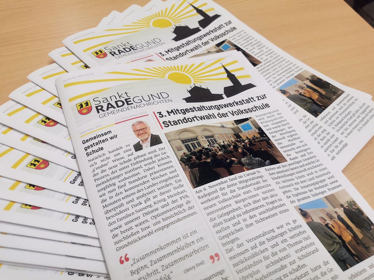 Gestapelte Zeitungen mit der Überschrift 'Sankt Radegund Gemeindenachrichten'. Eine zeigt einen Mann und diskutiert den 3. Platzierungsworkshop für die Volksschule. Ein Bild zeigt eine Menschenmenge in einem Saal.